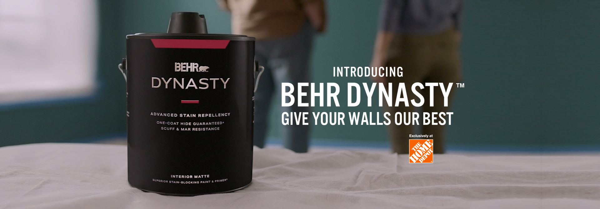 Couple getting ready to paint with a can of BEHR DYNASTY Flat interior paint and the words Introducing BEHR DYNASTY Give Your Walls Our Best in foreground.
