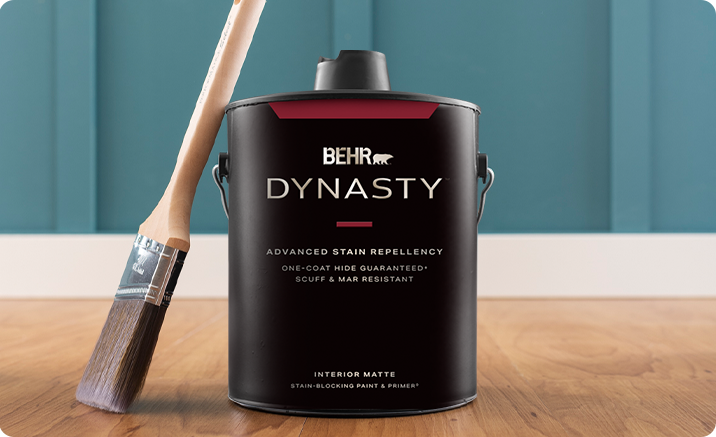 1 gallon can of BEHR DYNASTY Flat interior paint in front of painted wall with a paint brush propped up against the can