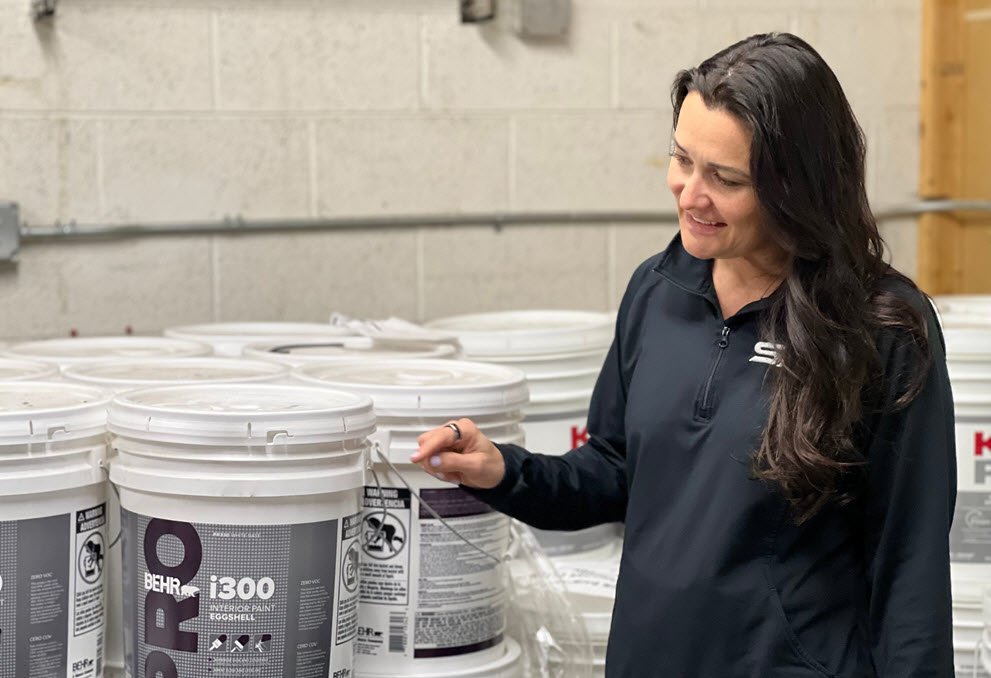 A woman in black shirt checking out a 5 Gallon of BEHR PRO Paint