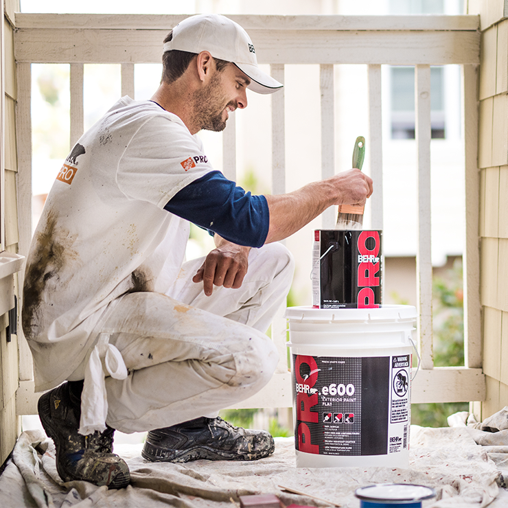 A close up shot of a BEHR PRO e600 1 gallon stacked on top of a 5 gallon. The body image of a contractor next to the can image shows him dipping paint brush on the 1 gallon paint can.