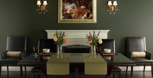 Stately traditional dining room with rich moss green on walls and white paint on fireplace mantle and trim. Leather dining chairs surround dark wood table.