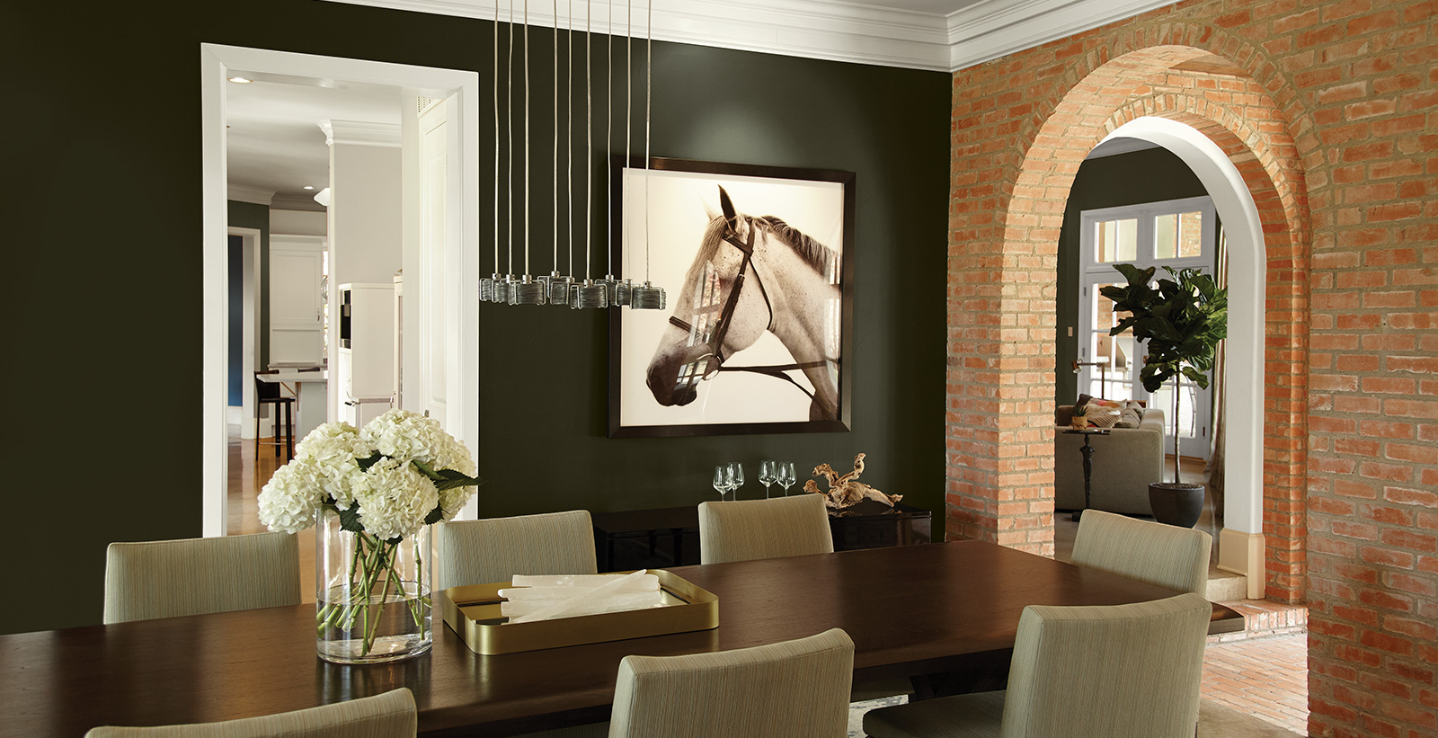 Contemporary farmhouse dining room with dark forest green on walls and white trim, brick accent wall surrounds an arched entryway.
