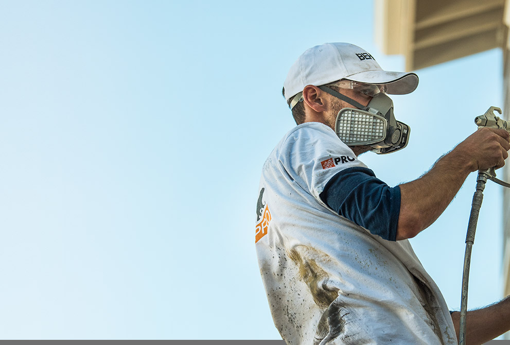 Image of a Pro Contractor wearing a hat and shirt with Behr logo spray painting an exterior wall of a house on a ladder.