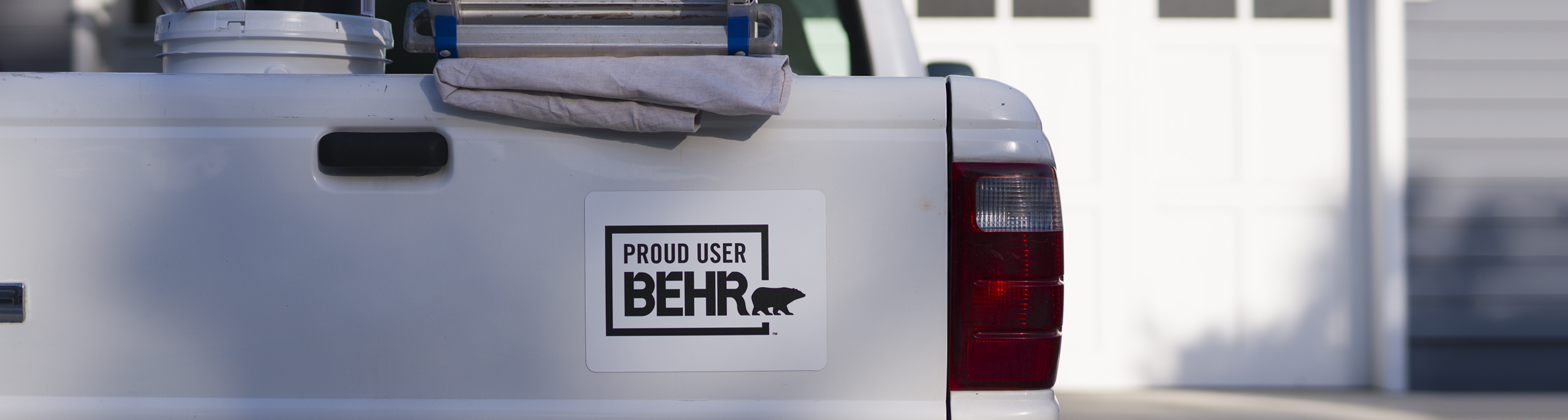 Desktop view of a pick up truck with a Behr sticker that says PROUD USER BEHR. On the back you can see a ladder, dropcloth, 1 gallon of BEHR PREMIUM PLUS Exterior Paint.
