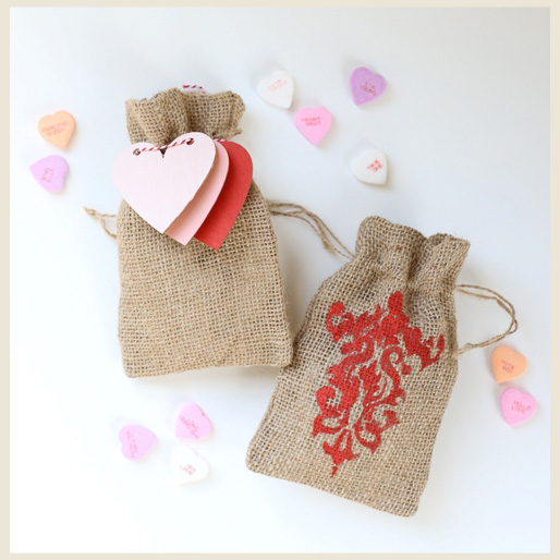 Two burlap bags decorated with red stencil stamp, paper hearts, and hard heart-shaped candies.