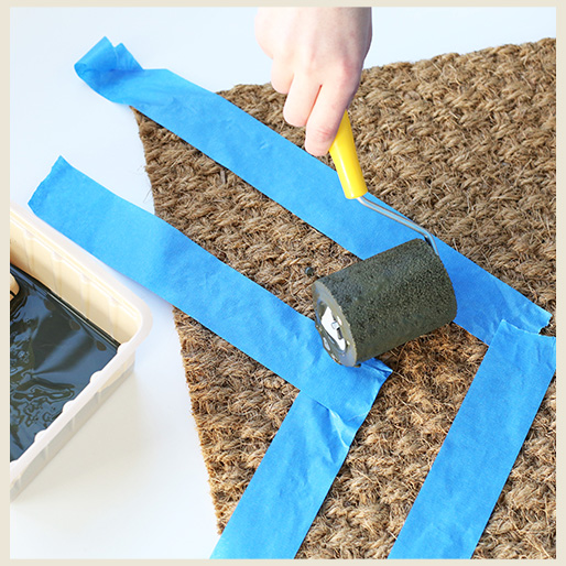 A frame showing a hand holding a roller brush with paint and applying onto jute doormat.