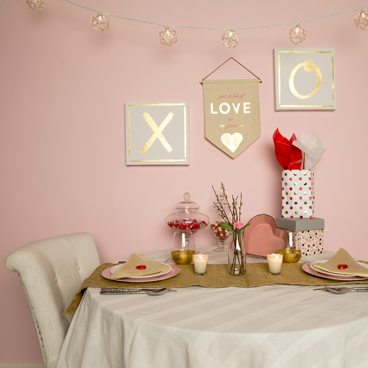 This image shows a creative way to set up a Valentines Day table setting. The wall is painted in Funhouse which is a muted pink. The wall has a X and O on canvas to symbolize hugs and kisses. There is a sign that says "put a lot of love in your heart" and also hanging overhead is small lights. 
The table setting has a large plate, small plate, napkin and two cups. 
There are chocolates on the table as well and two gifts. 
To finalize the look is a flower at the middle of the table and two candles. 