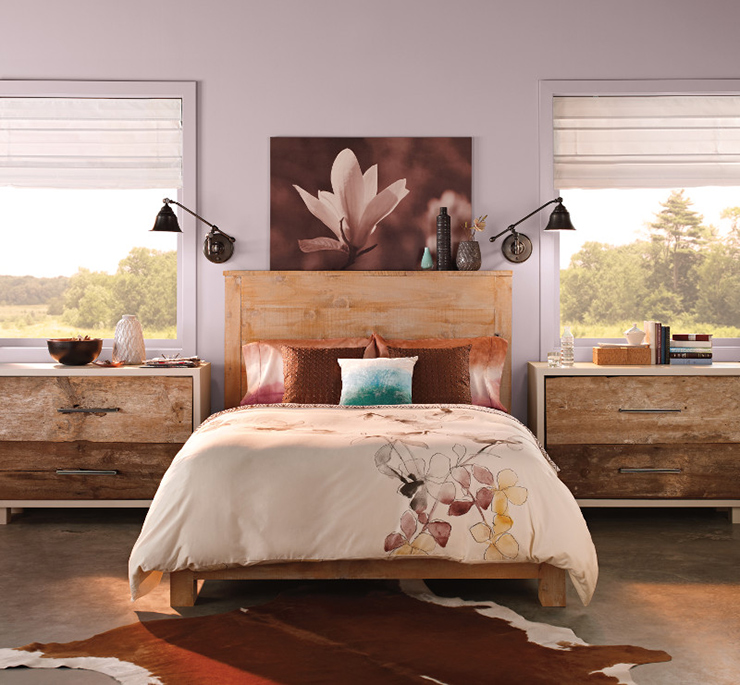 This is a country style rustic bedroom painted in Etiquette. There is a wooden headboard and wooden night stands. There is a portrait of a flower behind the headboard. The nightstands are decorated with books, vases, and a decorative box. 