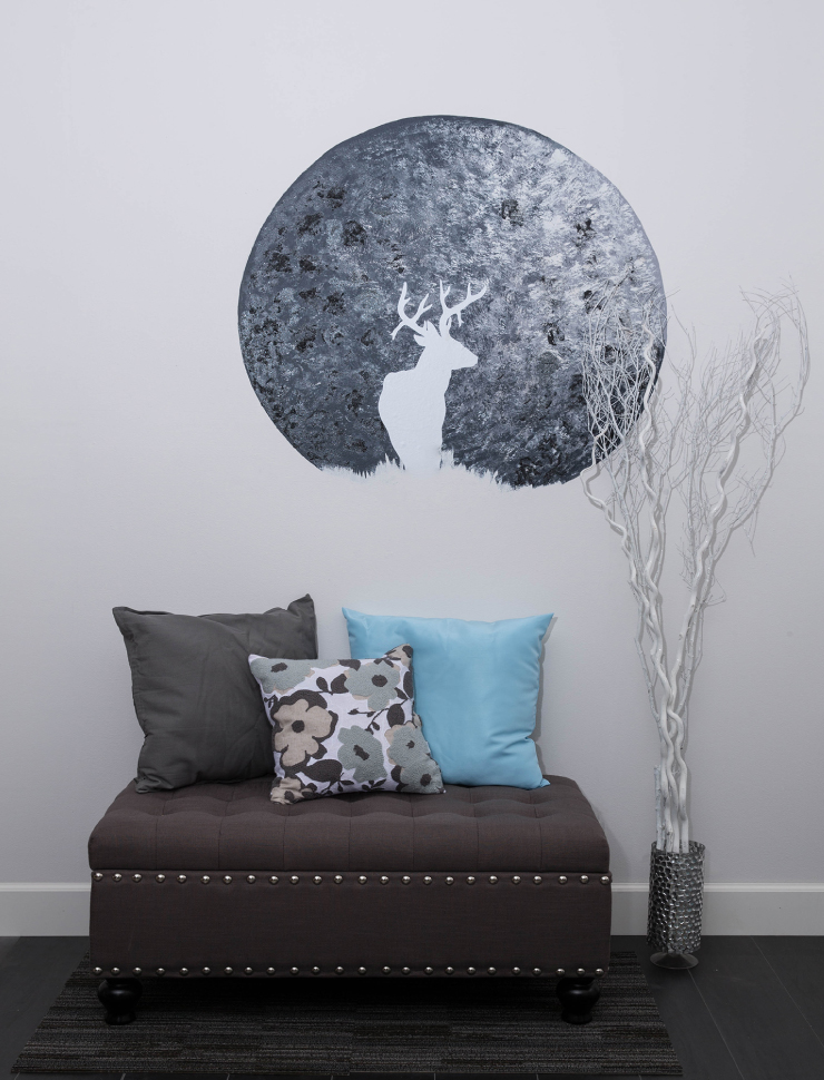A  sitting area with a brown ottoman and pillows. The wall behind it is painted a light gray with a painted moral of the moon.
