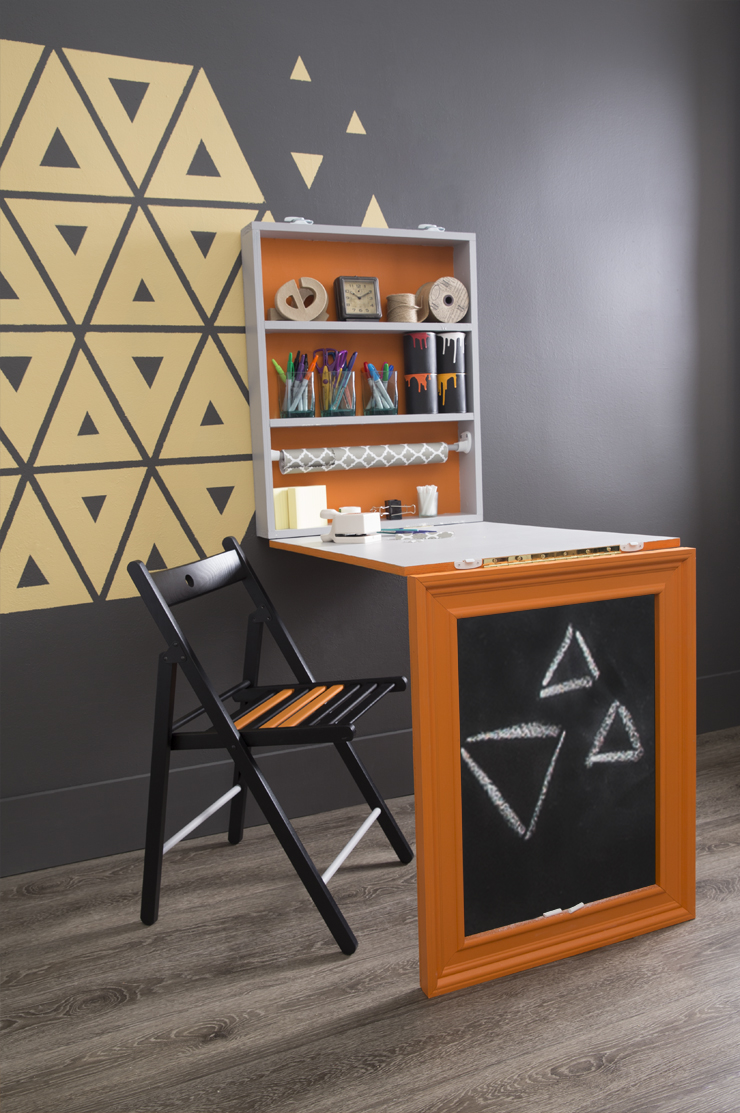 A room with a small desk and chair placed in front of the wall with the geometric shape painted on it.