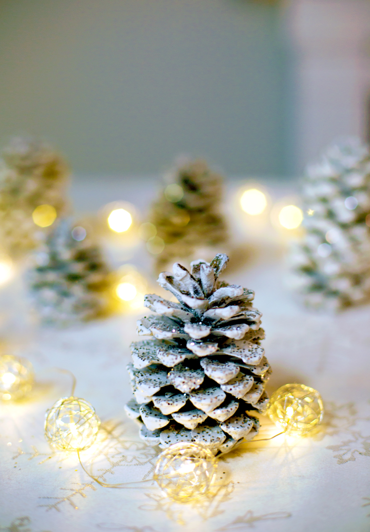 Glitter pinecones and lit gold string lights placed on a table.