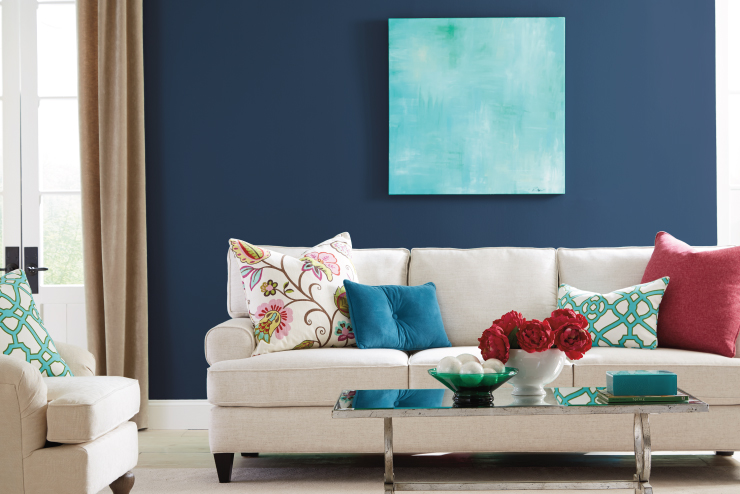 A one-wall living room featuring a dark blue paint color. A white sofa with solid and colorful, floral decorative pillows.