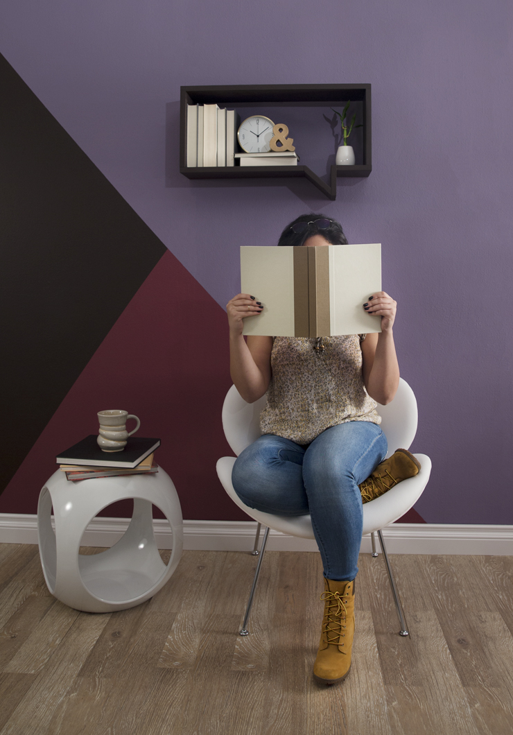 A person sitting on a chair with a book covering her face. The wall behind her is painted in geometric shapes using shades of purple. Above the girls is a book shelf shape like a thought quote.