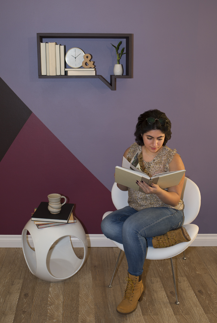 A person sitting on a chair reading a book. The wall behind her is painted in geometric shapes using shades of purple. Above the girls is a book shelf shape like a thought quote.