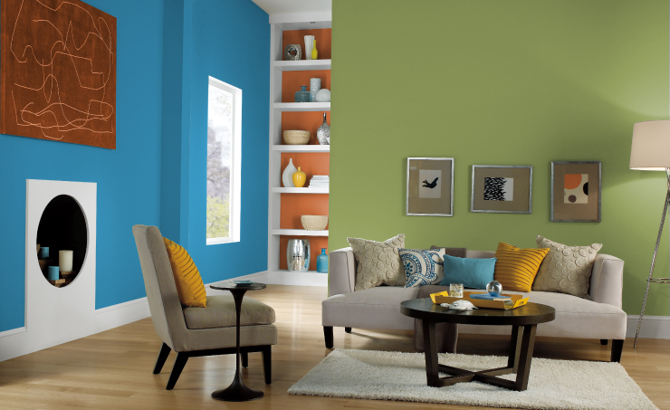 A living room painted in a green color. The room is open to a hallway painted blue and showing an orange and white bookshelf at the end.