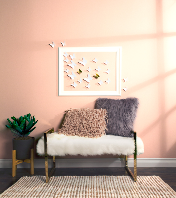A welcoming and youthful sitting area painted in a light color called Positively Pint. A white furry bench with metallic gold legs and shaggy decorative pillows. A pink wall decorated with a white frame and paper cut butterflies.
