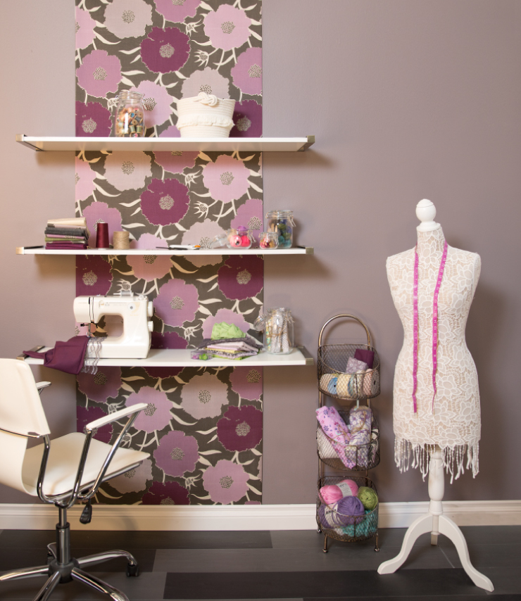 A sewing room with the wall painted in a purplish-gray color, Graylac. The wall has three shelves holding a sewing machine, material, spools of thread, jars with buttons. Behind the shelves is a strip of material that runs down the wall. The material has large purple and pink flowers on a dark gray background.