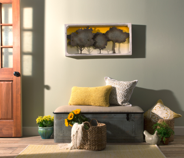 An entry with a green wall and a metal bench with yellow and cream pillows. The wall is painted a green color. On the wall is a hanging of  three metal trees sitting in front of a painted sunset sky.
