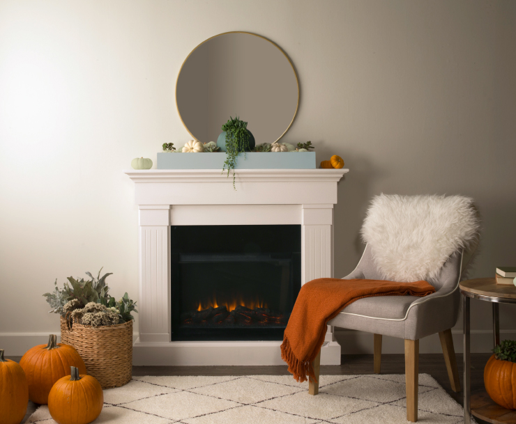 A lit fireplace. On the mantel is blue colored box holding pumpkins that were painted in white, blue, and green paint colors. 