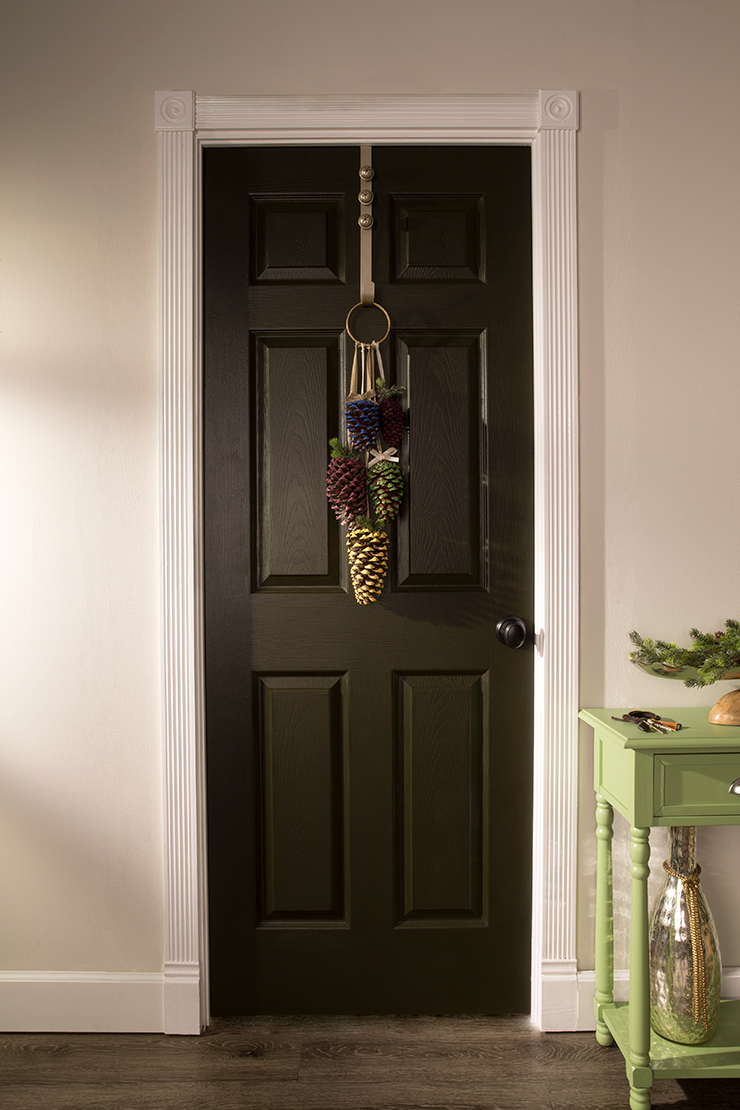 This image is of an interior door painted in Unplugged. 
It is decorated with painted pinecones hanging on the door with a door hanger. 
To the right of the door is a green console with keys peaking through and a branch of pine needles. 
Also a large vase on the bottom shelf of the console. 