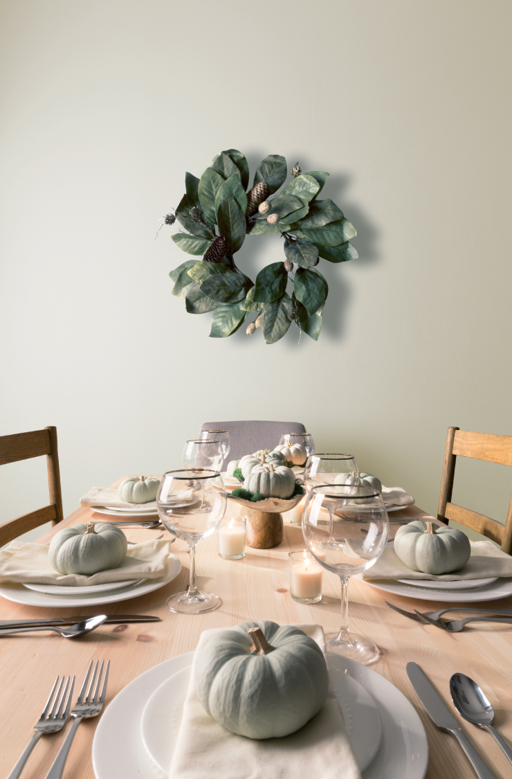 A natural wood dining table set in Thanksgiving décor of soft green colored pumpkins, white candles, white plates and cream color napkins. On the wall is a wreath made from large green leaves and pinecones. The wall is painted in Soft Focus.