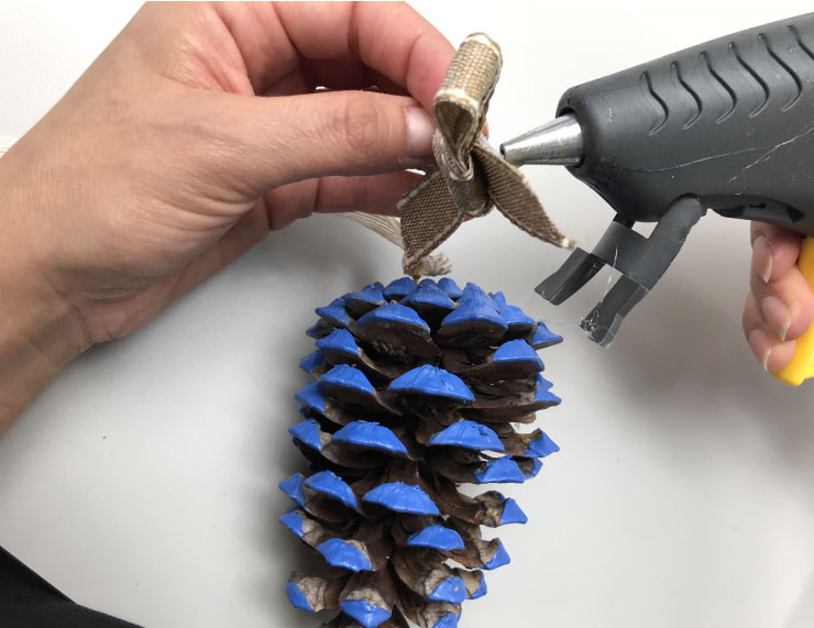 A bow being hot glued to the pinecone in order to hide the eye hook. 
