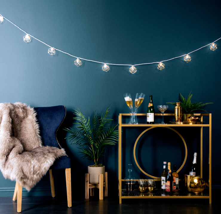 A sitting area with a deep blue velvet chair. In the chair is a dark cream colored soft furry blanket. Next to the chair is a liquor table with fun New Year’s Eve drinks. String lights hare hanging from the wall. Wall is painted in color Midnight in the Tropics.