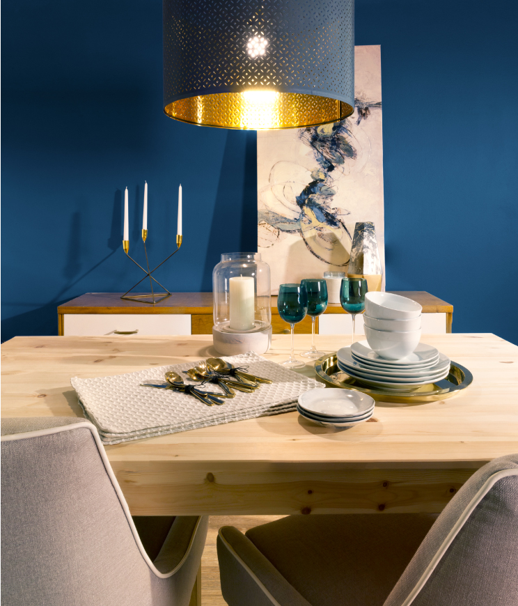A modern dining room painted in a deep blue color called Soul Search T18-14. Decorative elements include a white ceramic plates and metallic gold-ware, a modern lighting fixture and an abstract painting. 