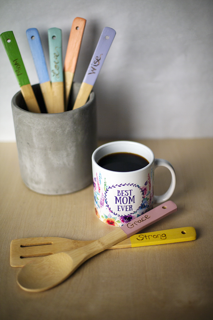 A counter showing a cup filled with coffee. The words “Best Mom Ever” if printed on the cup. Next to the cup is a ceramic container holding wood utensil. The end of each utensil is painted, and inspirational words are written on them; Grace, Strong, Wise, Love.