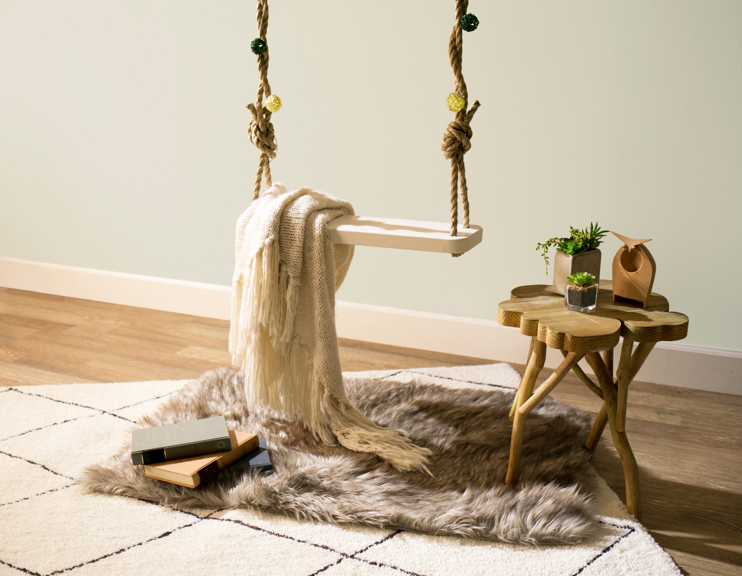 A tight crop of the swing and table next to swing with plants. Image also shows the floor with fluffy rugs and books.