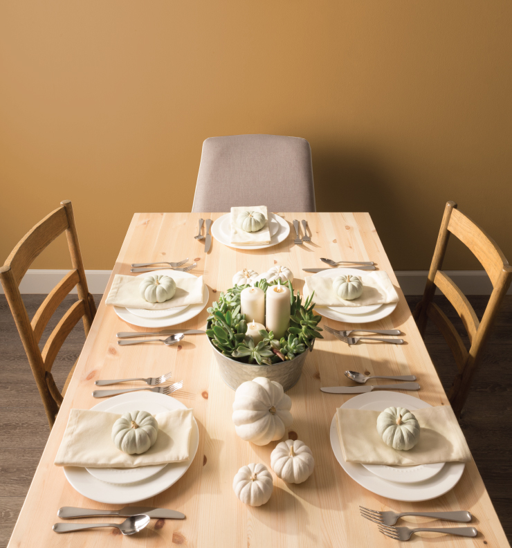 A wood dining table set for dinner with white plates and cream color napkins. The centerpiece is a tin vase with succulents and candles. The wall in the background is painted in Amber Autumn.