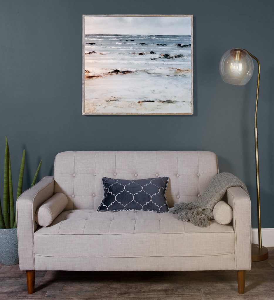 Living room showing a beige couch with a blue and white pillow and a gray blanket draped on the arm of the couch. The wall has a painting the ocean. The wall behind the couch is painted in Blue Metal.