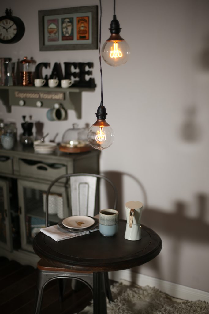 A close up of the small dining area with a small table and chair with hanging lights above. The table is set with a coffee mug, plate and pot of pressed coffee.