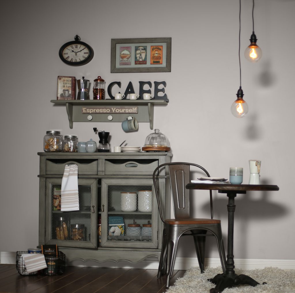 A small dining area with the wall painted in Cotton Grey. There is a small table and chair with hanging lights above. Against the wall is a buffet cabinet displaying coffee items; mugs, grinder, café sign.
