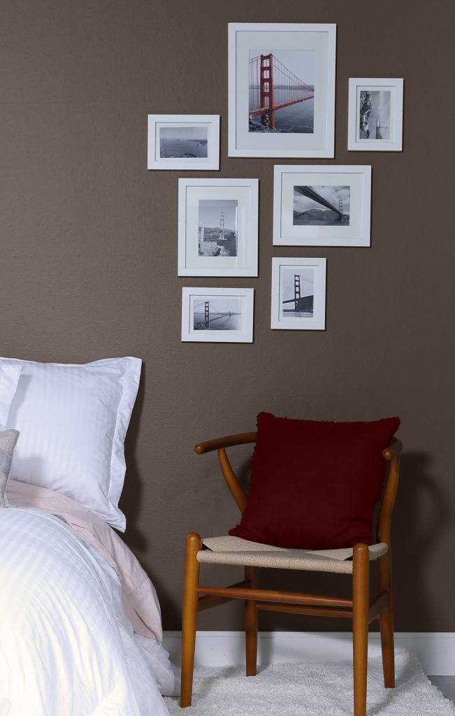 A bedroom featuring the color  KINDLING N200-6. The wall is decorated with white frames and the room has a wooden chair and a bed with a white comforter. 