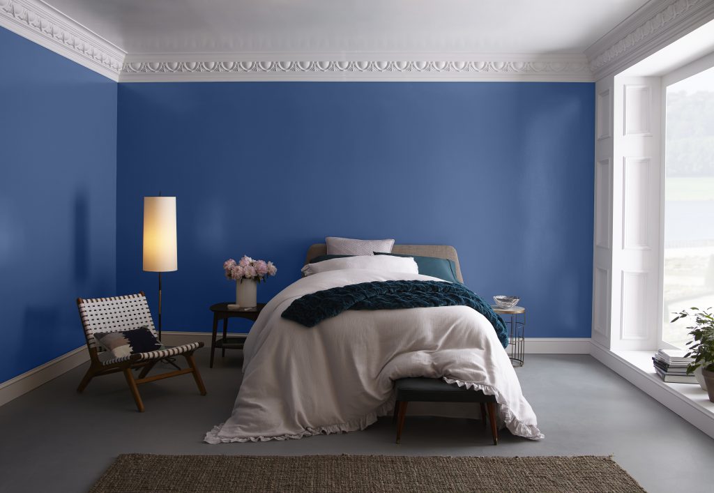 A blue bedroom showcasing large wall-sized window and fluffy, comfy bed. 
White painted woodwork including ornate crown molding. 