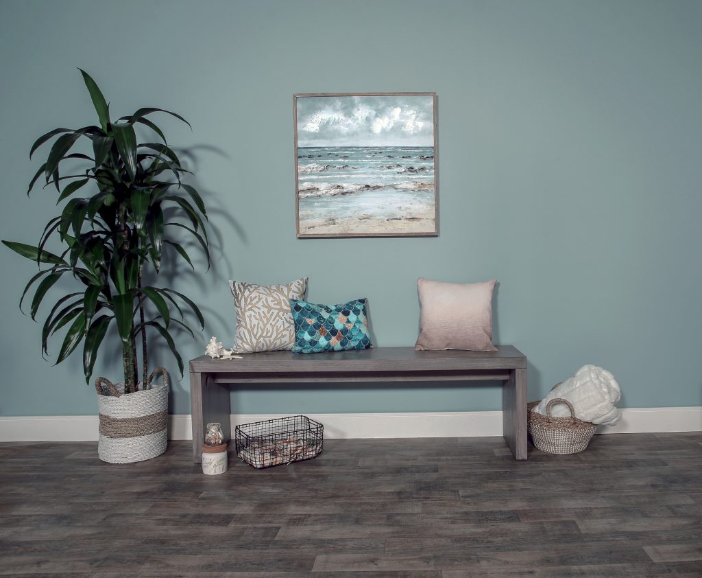 This is a coastal inspired entryway. The wall is painted in Watery which is a dusty shade of blue with a subtle green undertone. 
There is a wooden bench with aquatic pillows, a basket of seashells and a basket with a cozy blanket. 
To finish it off there is a portrait of the ocean hanging on the wall and also a glistening green palm tree. 