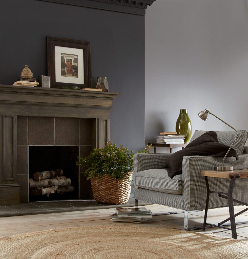 A living room featuring a fireplace with an antiqued wood finish mantle, the wall is painted in dark gray color called Graphic Charcoal. A mix of decorative elements including stacks of books, tin box, wall artwork, ceramic vases and plant in a basket.