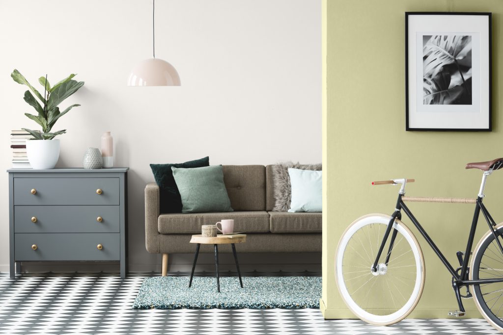 An apartment living room with an accent wall painted in green color called Back to Nature. A smoky teal cabinet decorated with potted fiddle fig plant, ceramic vases, a stack of books and metallic gold drawer pulls. A sofa with several natural and green toned decorative pillows. 