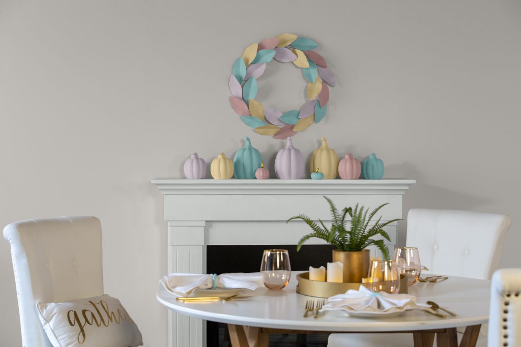 A dining area painted in an off-white color.  All white dining furniture and wood fireplace mantle.  A fireplace mantle decorated with painted ceramic pumpkins and metal wreath on the wall. 