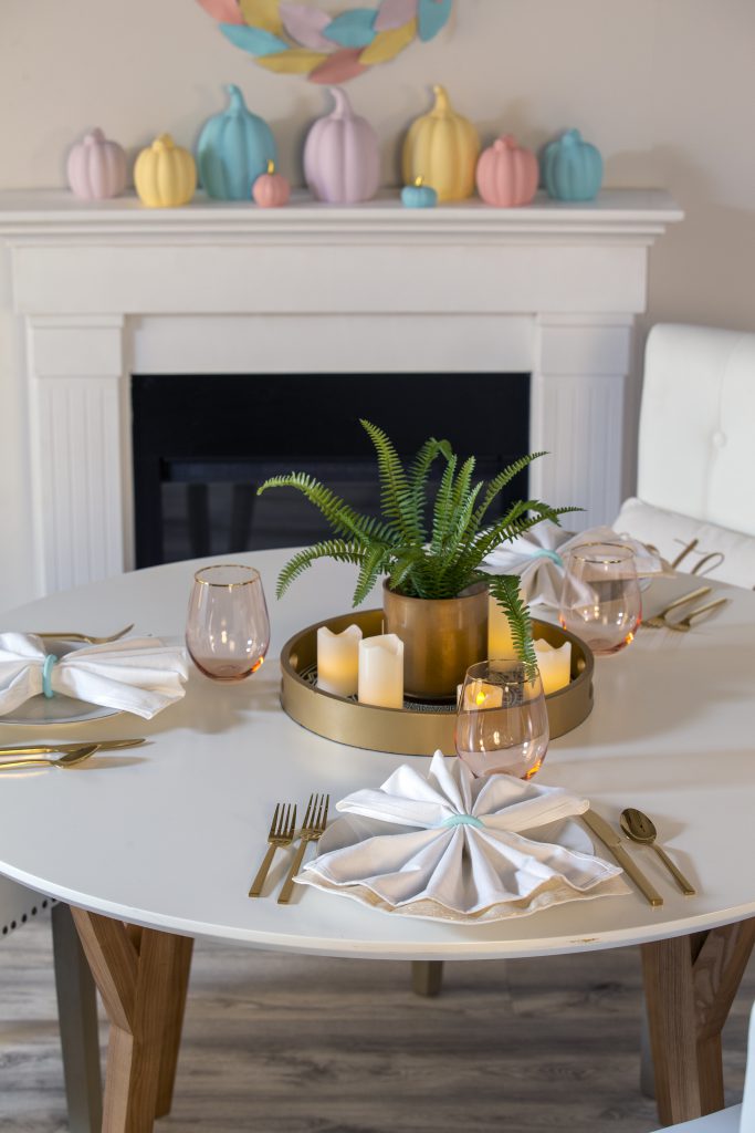 A simple table setting for three featuring gold eating utensils.  A fireplace mantle featuring ceramic pumpkins painted in pastel colors and metallic gold touches. 