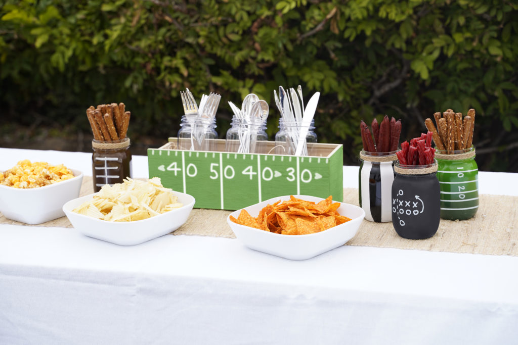 Buffet table set with snack placed in mason jars that were painted in a football theme. The four mason jars resemble: football, referee, score board, football field. A box is in center holding plastic wear. The box is painted to resemble a football field.