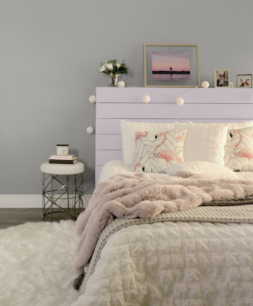 Bedroom featuring a wood plank headboard paint in Dusty Lilac. The bed has white bedding, with pink, gray and white blankets, and flamingo pillows. Large rose ball lights wrapped around the headboard. 