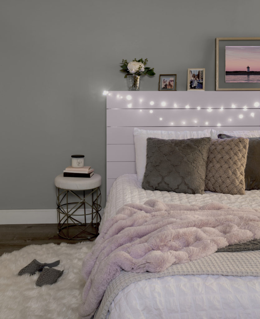 Bedroom featuring a wood plank headboard paint in Dusty Lilac. The bed has white bedding, with pink, gray and white blankets, and solid gray and brown pillows. Small lit twinkle lights wrapped around the headboard. 