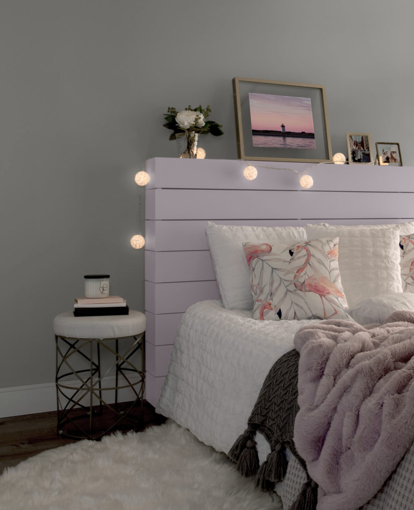 A side angle of a bedroom featuring a wood plank headboard paint in Dusty Lilac. The bed has white bedding, with pink, gray and white blankets, and flamingo pillows. Large lit rose ball lights wrapped around the headboard. 