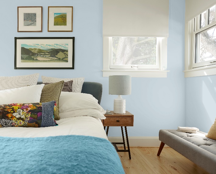 Bedroom painted in light blue color called Light Drizzle.  The bed is dressed with layered bedding and multiple pillows.   The rest of the bedroom is decorated with a side table, a lamp, wall artwork, a comfy sitting bench.  The overall feeling of the room in very light and airy as there are two windows that allow for plenty of daylight. 