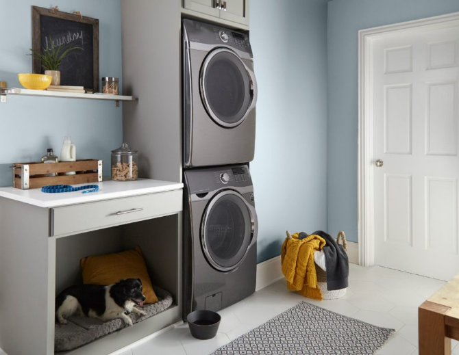 A laundry room with gray finish stack-able washer-dryer.  the room features a pet sleeping and eating area.  The walls are painted in light blue color called Light Drizzle. 