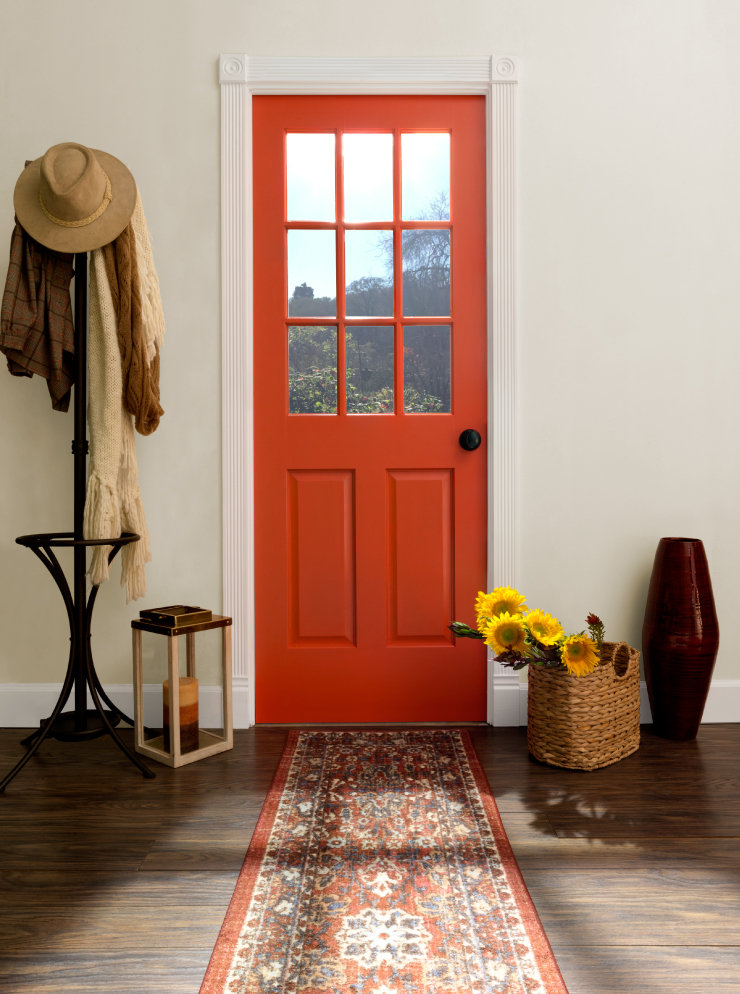 Accent interior door painted in Spirit Warrior. To the left of the door is a coat hanger and a lantern with a candle. To the right of the door is a basket with fresh sunflowers and a large standing vase. To the middle of the door is a runway carpet. 