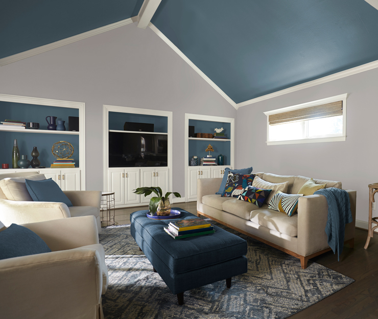 This image shows a vaulted accent ceiling in Blueprint. It is a living room with sofas embellished with pillows and a blanket. 
There is a long blue ottoman in between the sofas.             
Also against the back wall is a built in wall unit also painted in Blueprint as an accent.                                                                                                                            