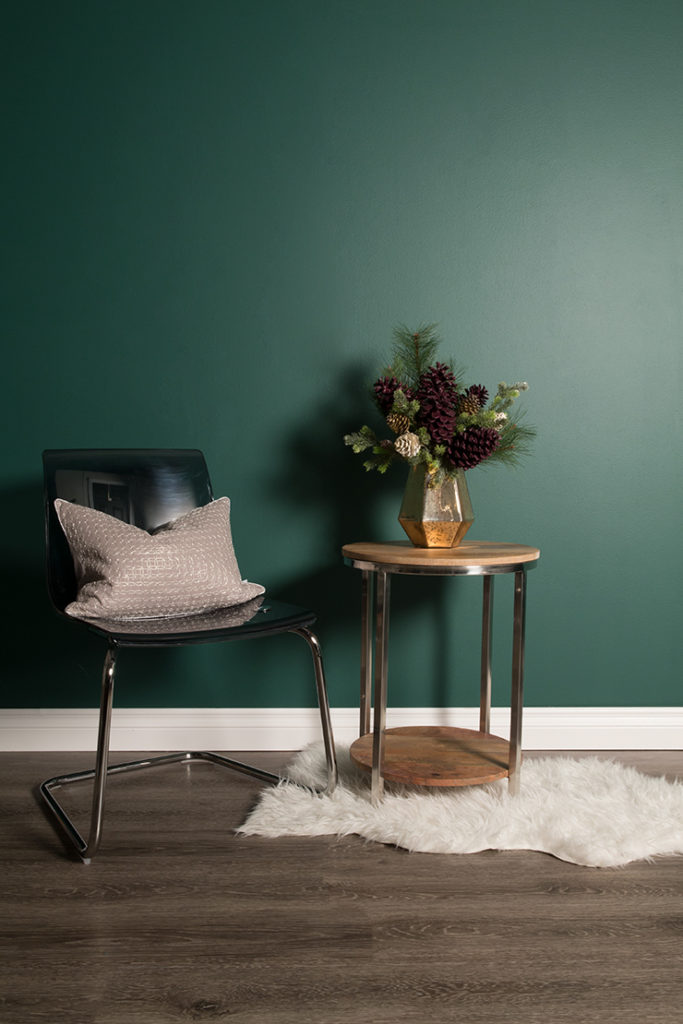 This image is a seating area with a wall painted in Equilibrium. There is a chair with a pillow and to the right is a wooden table with the DIY painted pinecones in a bouquet. 